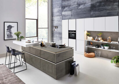 Designküche von Nolte mit weißen Hochschränken und grauer Kochinsel in Schieferoptik mit Kehlleistenbeleuchtung.