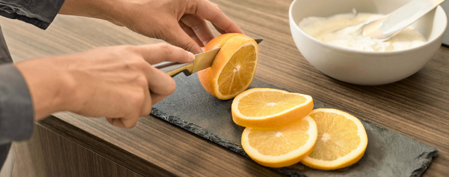 Küche von Nolte – Detailaufnahme beim Schneiden einer Orange auf dunkler Arbeitsplatte, modernes Küchenambiente mit natürlichen Materialien